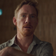 A man in a beige shirt in front of a brown background looks at something with curiosity.